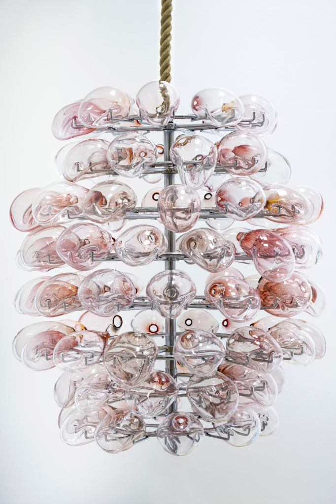 A metallic rack suspended from the ceiling by a rope, adorned with various drooping pale pink glass forms forming a large oval construction