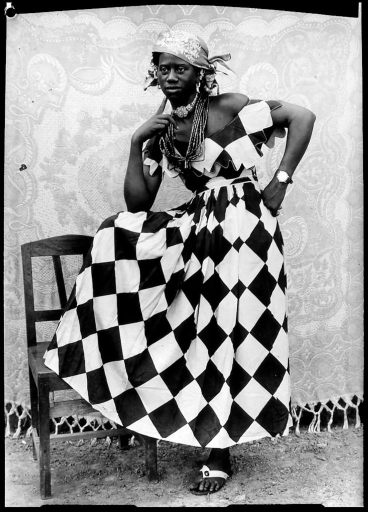 Black and white photo of a young Black women in checker patterned dress posing with her leg up on a chair, left hand on hip, and right hand up to her chin