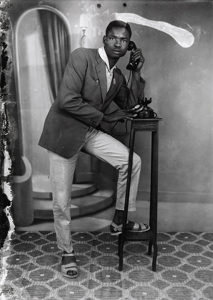 Black and white photo of a young Black man in a suit posing with a rotary phone and his leg up on a pedestal step