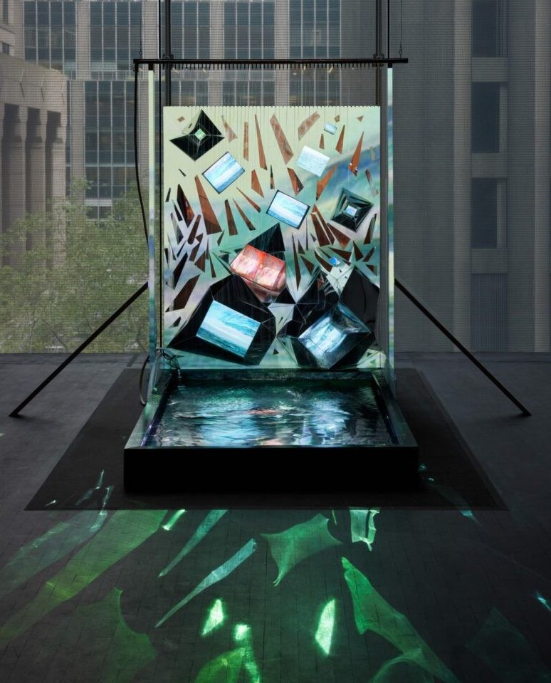 Sculpture of a large upright panel with several inlaid tv screens, standing over a shallow rectangular pool of water reflecting the videos