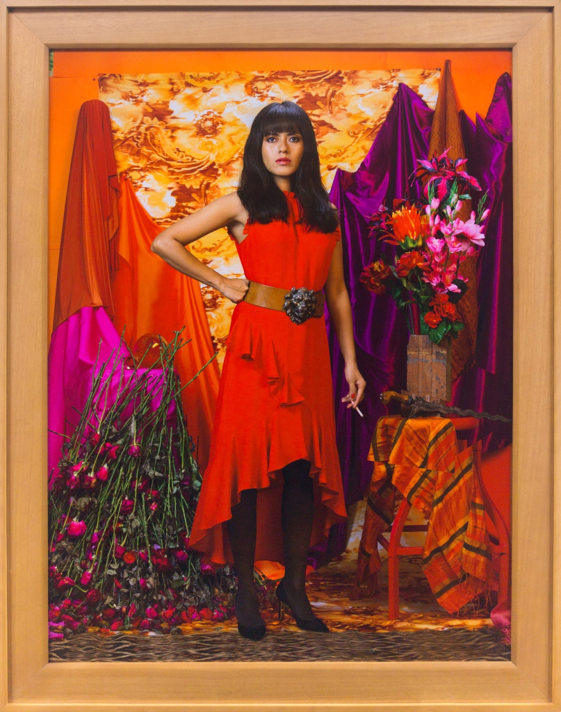 Photo of a young woman with long brown hair in a red dress, standing within a red, orange, and purple environment of draped fabrics and flowers, holding a cigarette in one hand