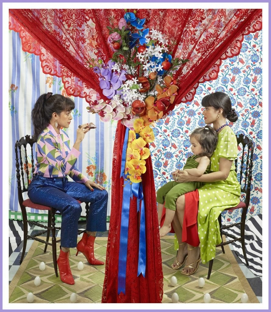 Photo of a young woman with long brown hair sitting across from her double holding a toddler in her lap. Both sit in wooden chairs in patterned rooms in a mirrored composition, with a lacy red curtain decorated with flowers draped in the center, separating the two spaces