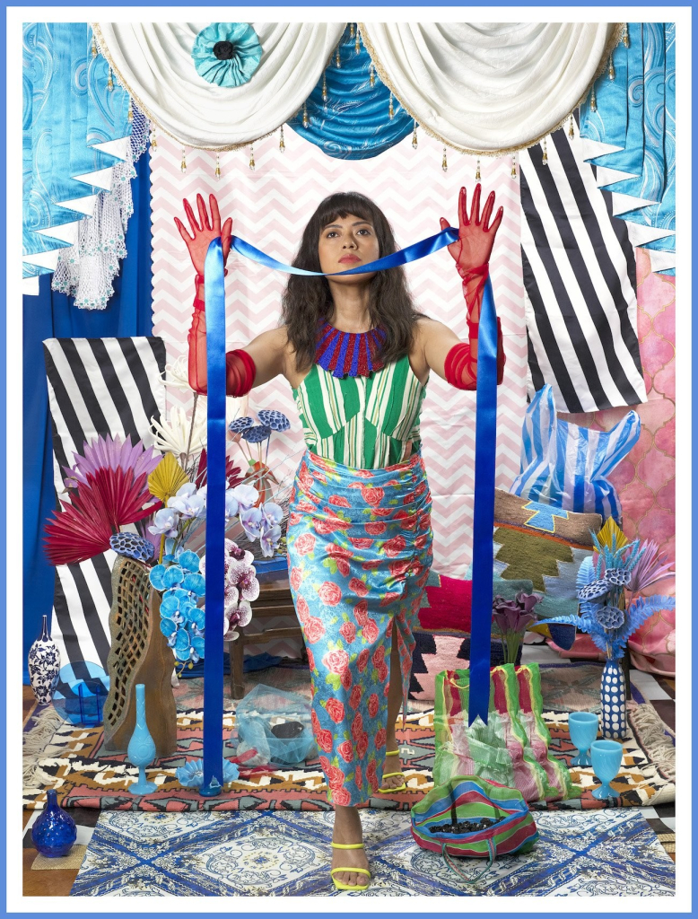 Photo of a young woman with long brown hair standing in a room decorated with patterned fabrics, colorful ceramicware, and flowers. She is dressed in a multi-patterned outfit and holding a long blue ribbon in both hands, leaning forward on one leg.