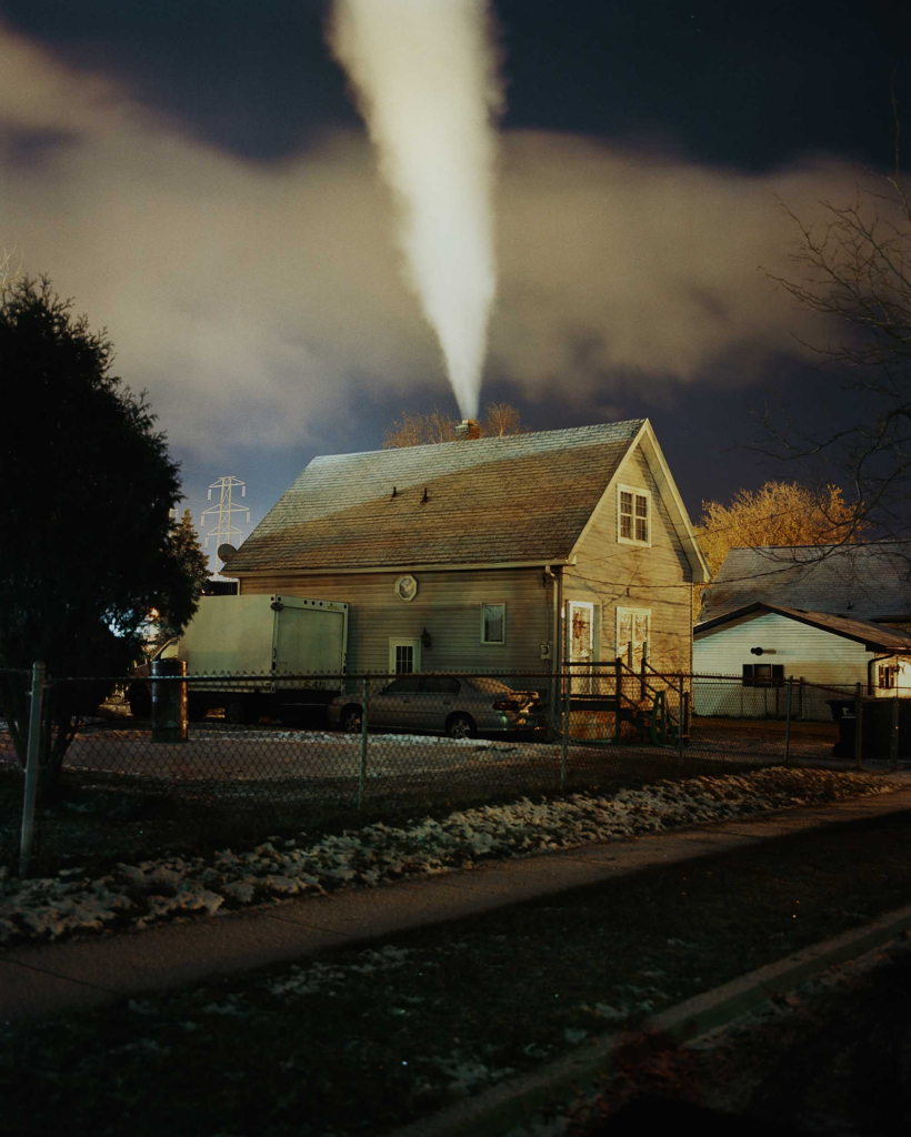 Vertical photo of a small house at dusk in a quiet residential neighborhood with some snow on the ground, and a white stream of smoke bursting through its chimney