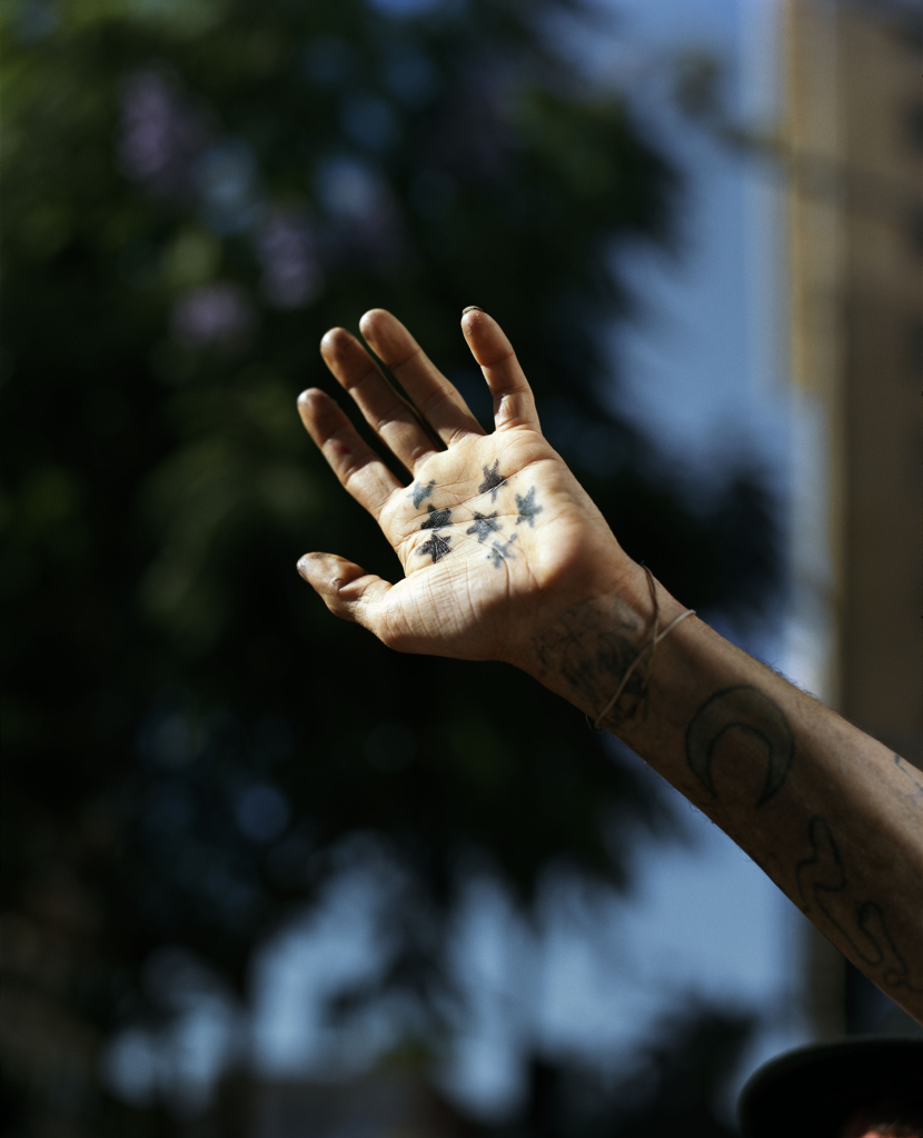 Vertical photo of a hand outstretched to sunlight, palm out covered with star tattoos and other ink running down the arm, a blurred tree in the background