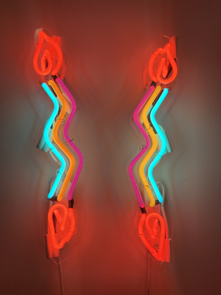 Neon wall sculpture of two wavy vertical shapes mirroring each other, with red candle flames at their tips