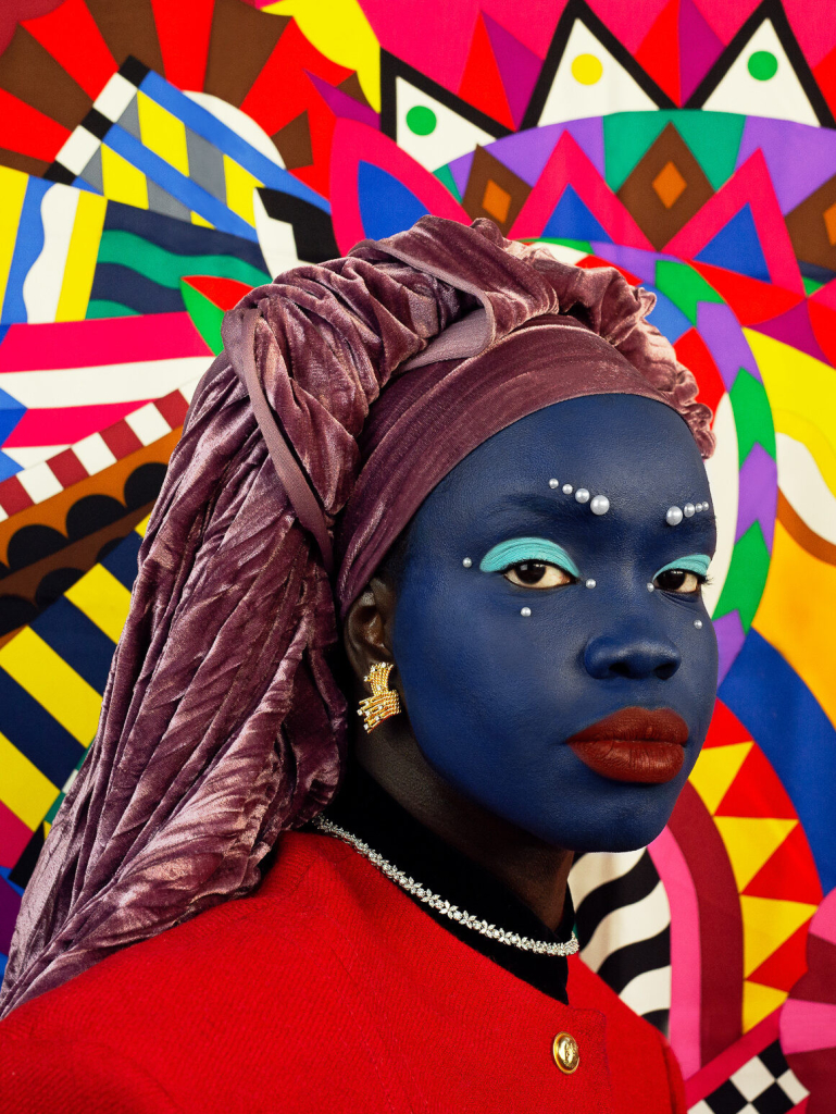 Photo portrait of a young Black woman with her face painted blue wearing a velvet purple headwrap, against a brightly colored geometric pattern background