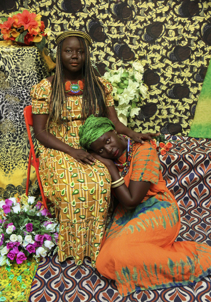 Photo of two seated Black women looking at the camera, one curled up on the floor with her head in the lap of the other. Both are wearing long patterned dresses, flowers and colorful patterned textiles arranged as the backdrop