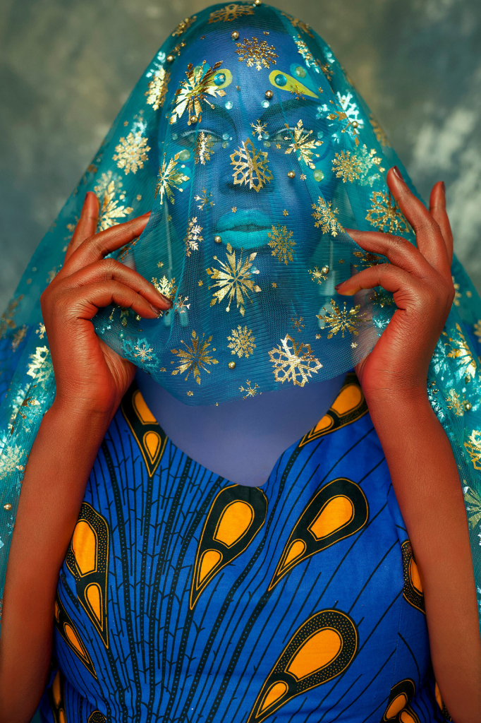 Photo of a young Black woman with blue skin makeup holding a blue and gold patterned veil over her head and face, wearing a blue and yellow patterned textile