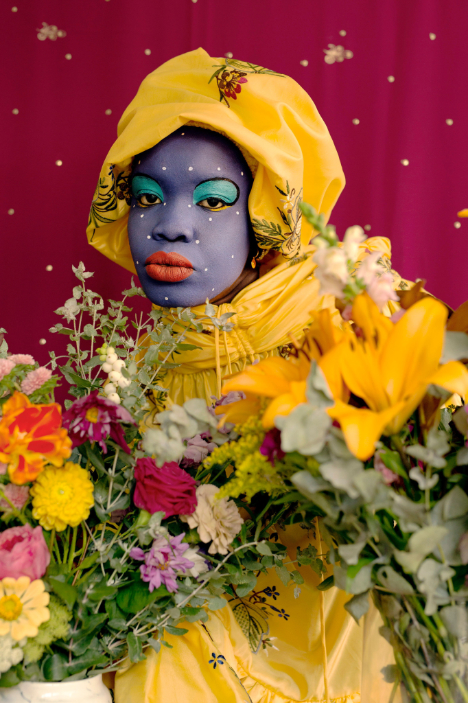 Photo of a young Black woman with blue skin makeup wearing a yellow headwrap and dress, with bouquets of flowers arranged in front of her torso and a magenta and gold textile hanging behind her