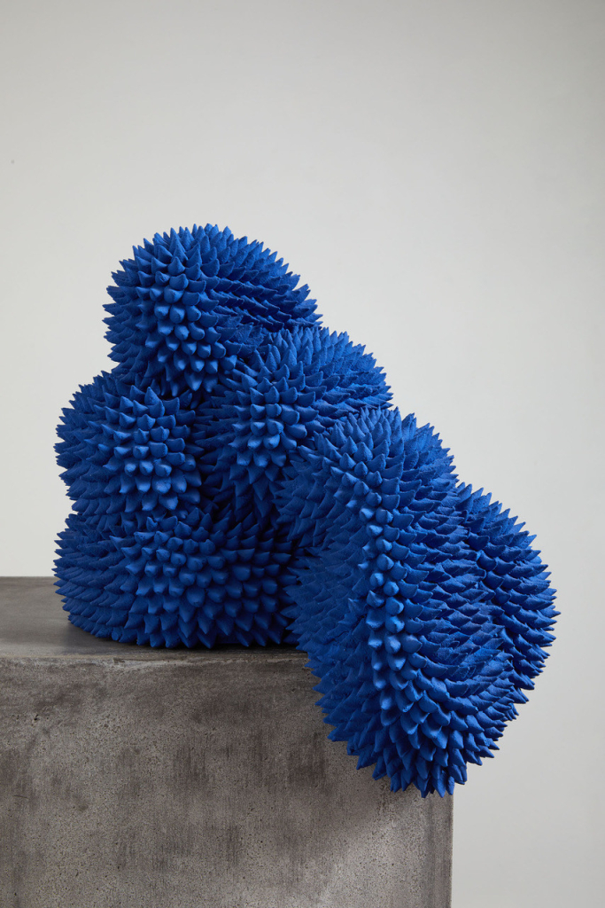 Abstract sculpture of a bright blue biomorphic shape covered in prickly points, sitting atop a concrete block