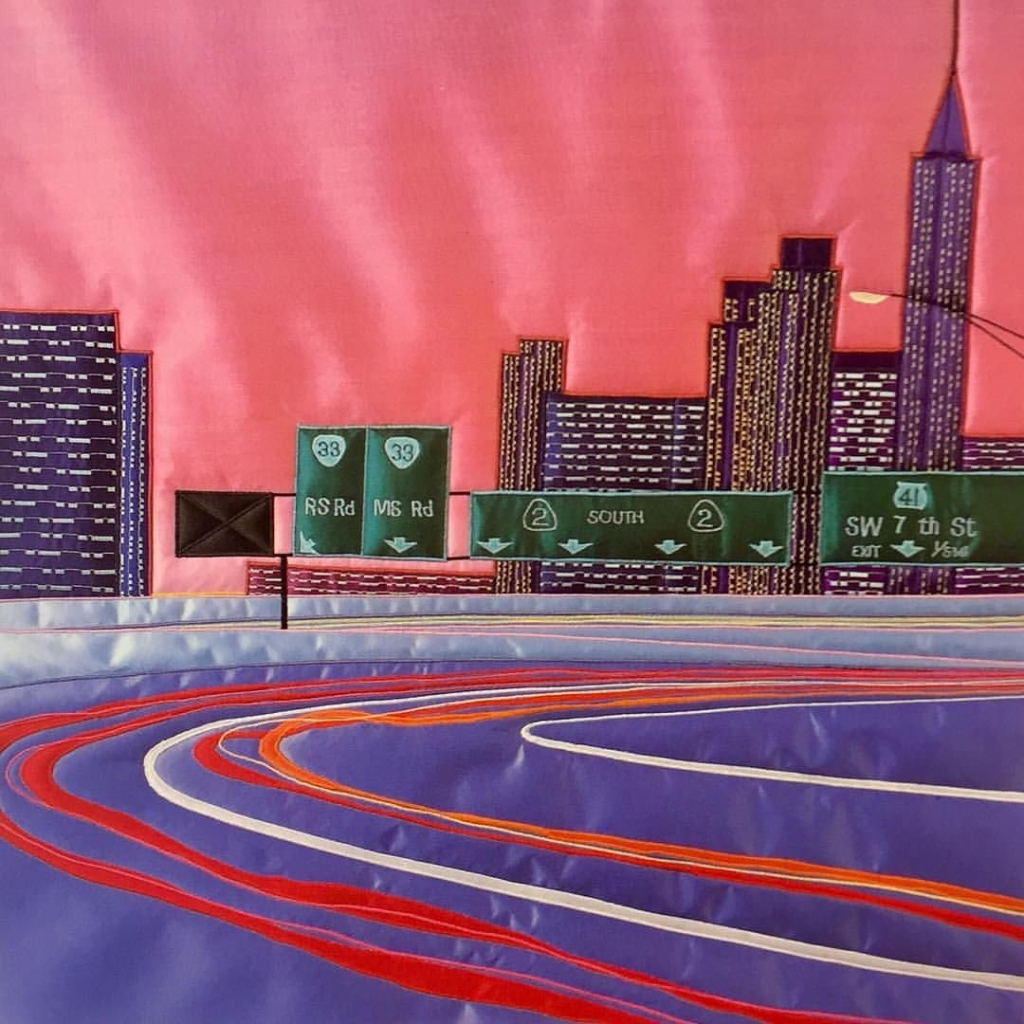 Embroidered scene on silk fabric, showing a city scene with purple and red road curving in the foreground, with purple skyscrapers lit up with lights and a pink background