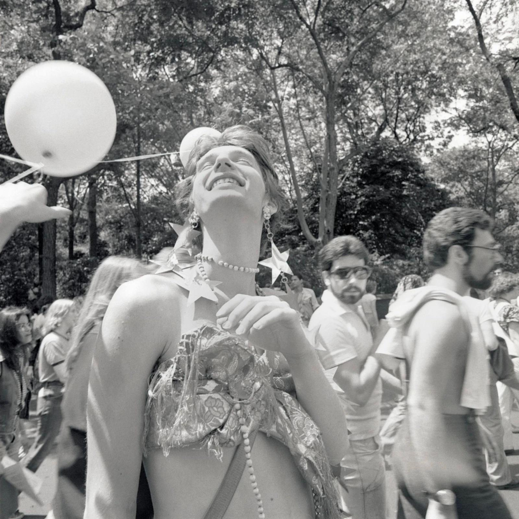 Black and white photo of a person wearing a shiny crop top and metallic star earrings, their head thrown back with a big smile and a crowd of other marchers behind them