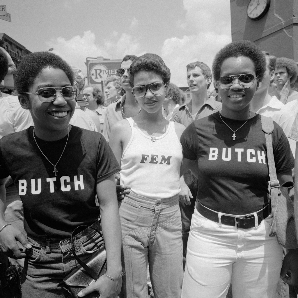 Black and white photo of a three young Black women posing and smiling with shirts reading "Butch" and "Fem"