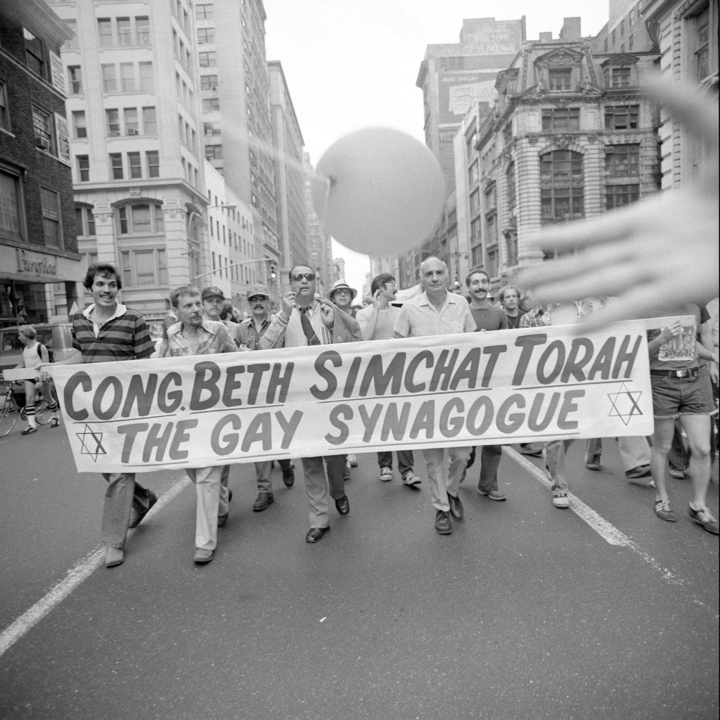 Black and white photo of a group of men marching and carrying a banner reading "Cong Beth Simchat Torah The Gay Synagogue"