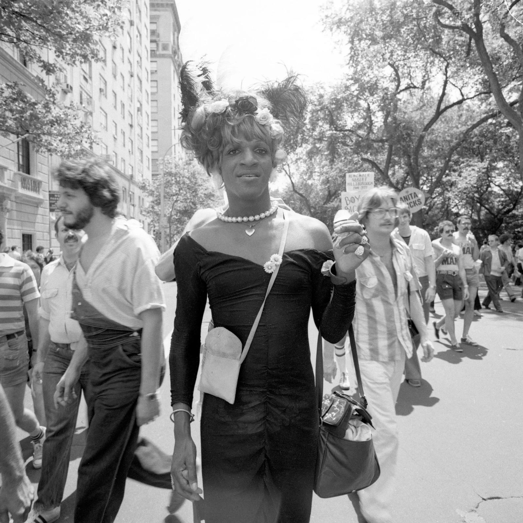 Black and white photo of a Marsha P Johnson walking towards the camera in a black dress with various folks behind her marching with signs