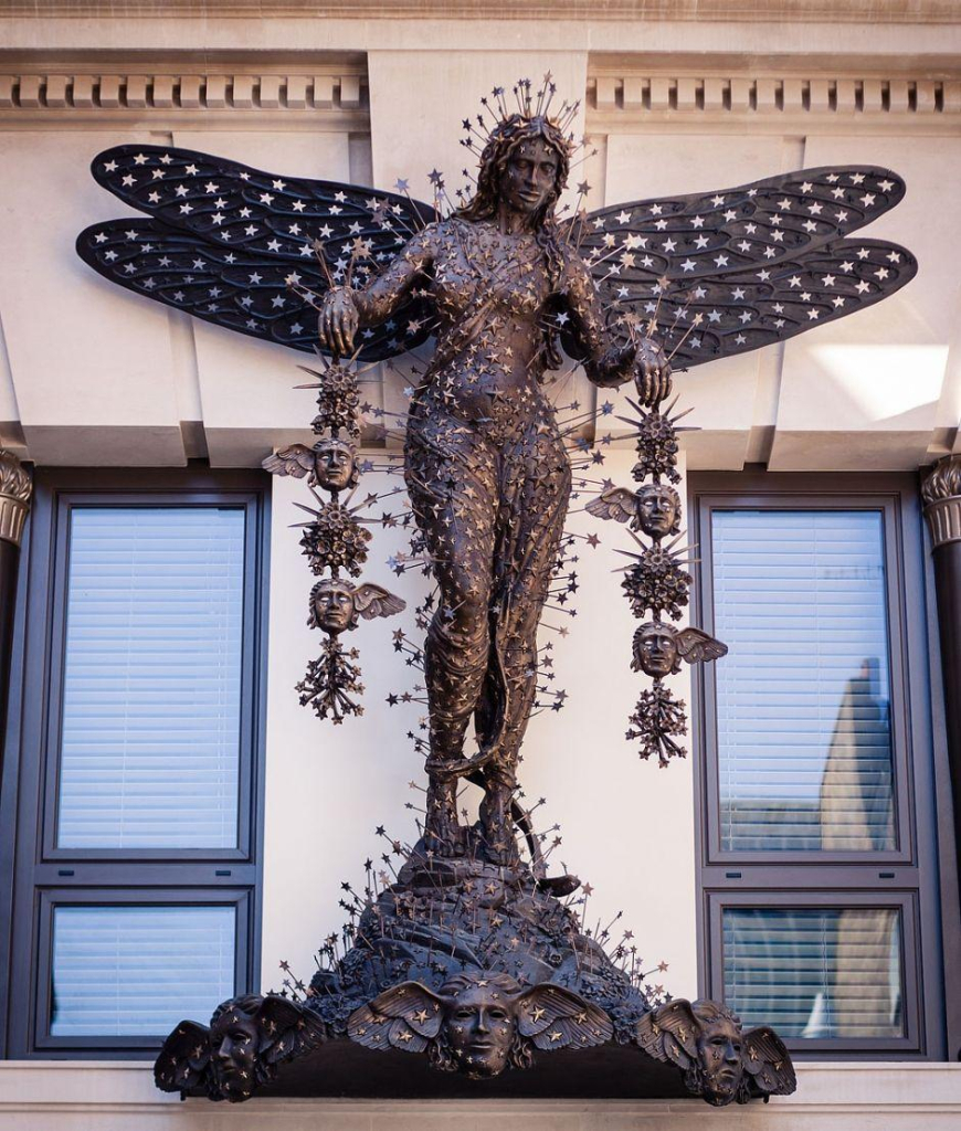 Sculpture of a nude woman covered in stars with dragonfly wings, holding lines of masks from her hands