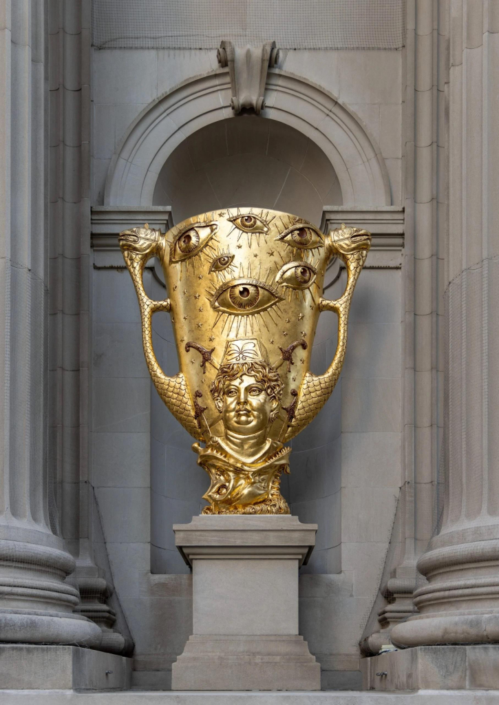 Gold sculpture of a large two-handled vase engraved with large floating eyes and a portrait of a puffy-cheeked white man at the bottom