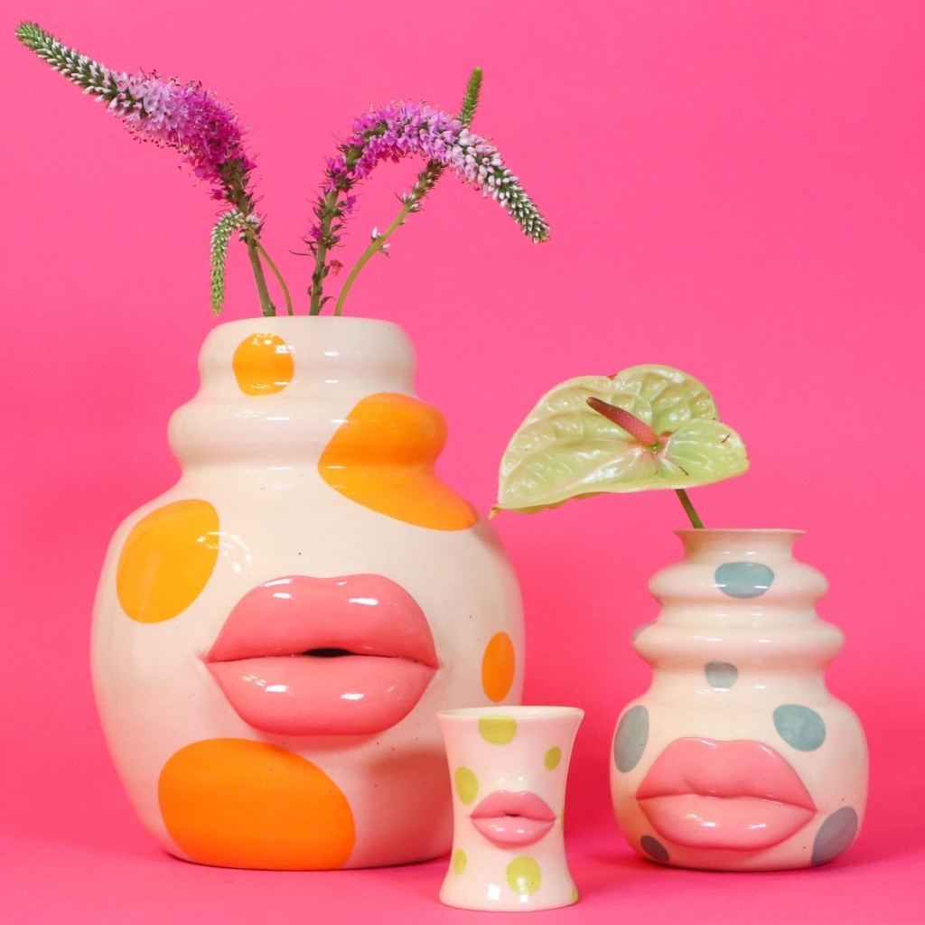 Two ceramic vases and a small cup, all with plump pink lips at their centers and colorful painted dots, sitting against a pink background