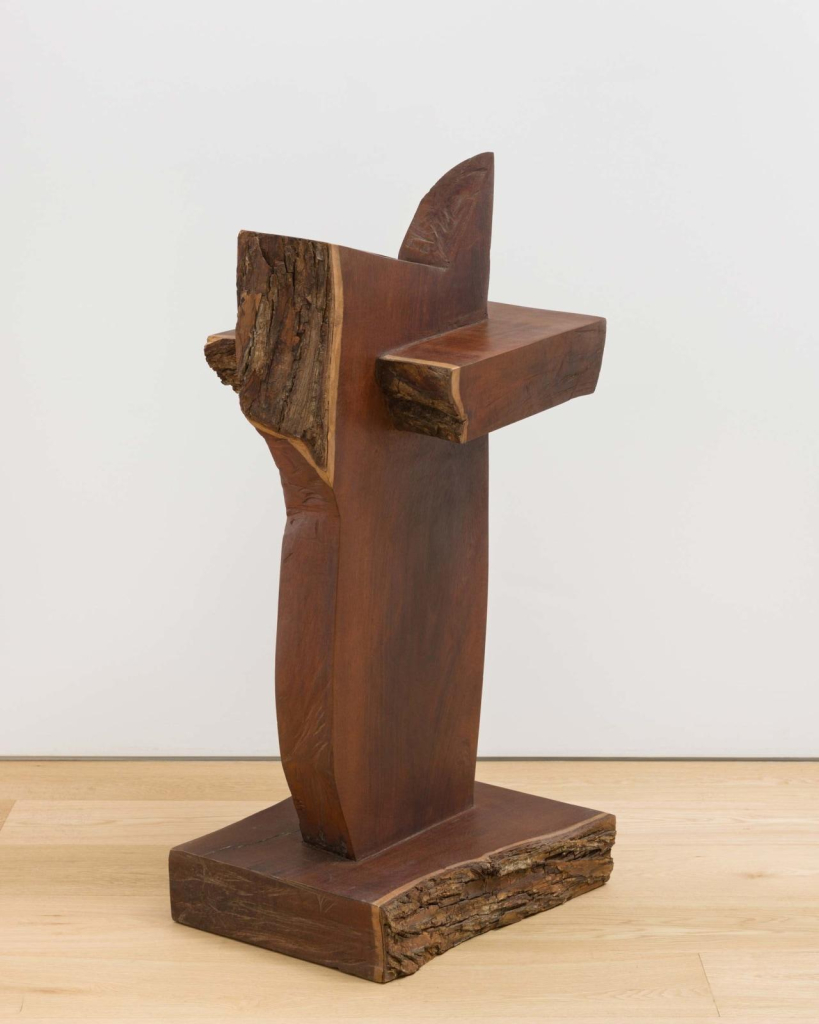 Abstract wooden sculpture of a smooth central vertical shape on a square base, bisected by a smaller rectangular shape near the top, with some sections showing jagged edges