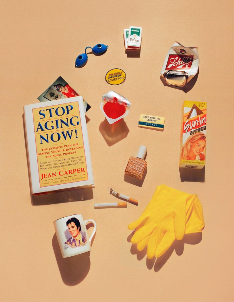 Still life photo of a collection of items arranged on an orange cream background, including a book "Stop Aging Now!", an Elvis mug, and cigarettes