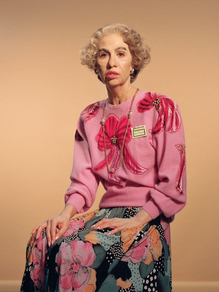 Portrait photo of the artist dressed as an older woman wearing a floral pink sweatshirt and long floral skirt, sitting on a stool against an orange cream background