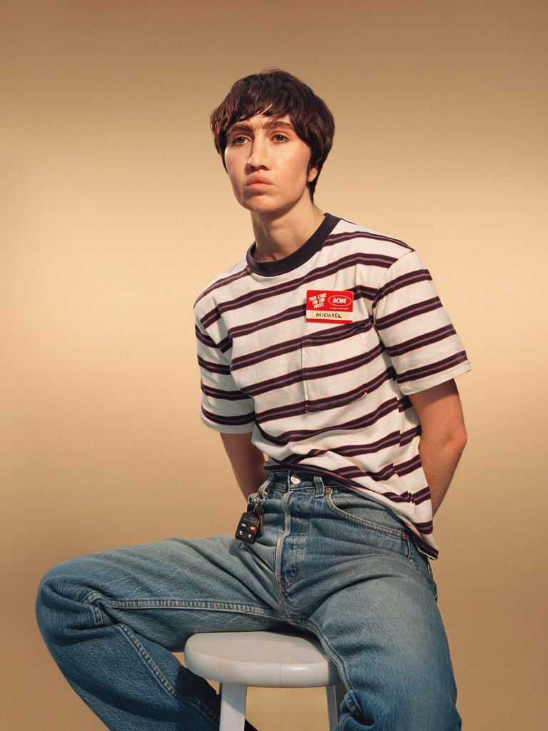 Portrait photo of the artist dressed as a young man in a striped tshirt and jeans, sitting on a stool against an orange cream background