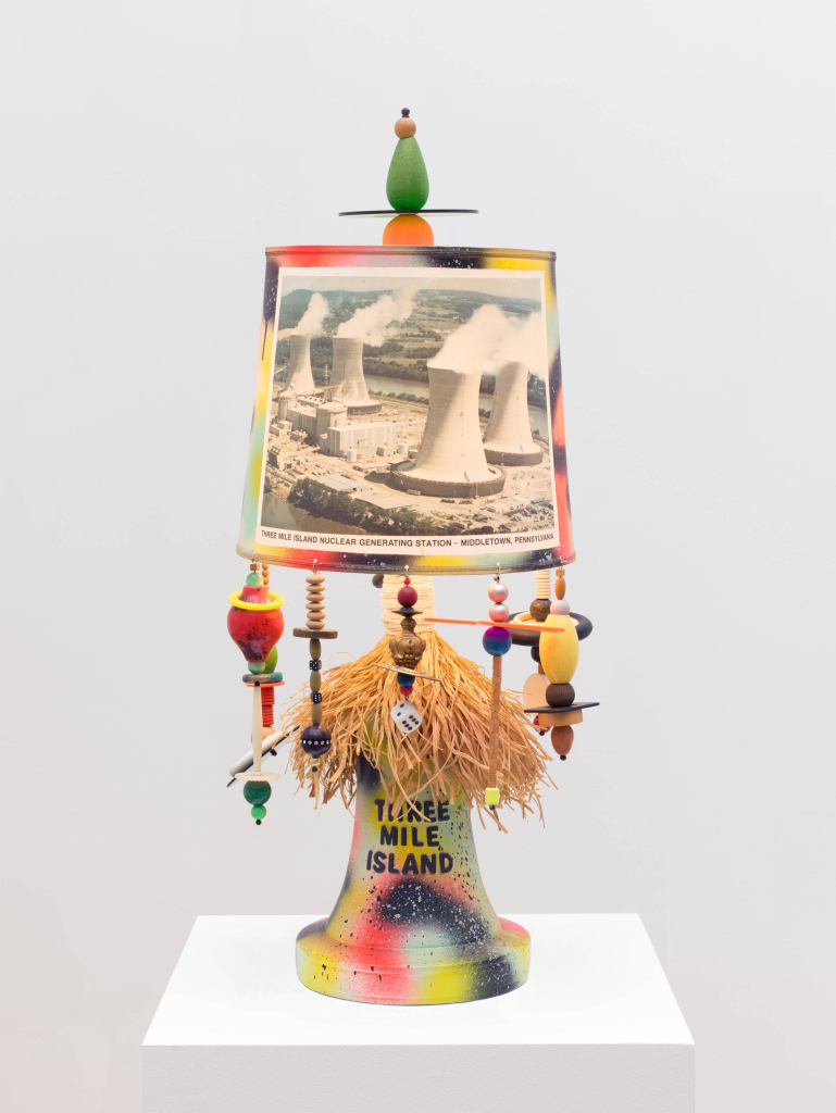 Table lamp spray painted in bright colors, with a photo of a nuclear power plant on the lamp shade and the words "Three Mile Island" printed on the base. Various abstract shapes dangle from the shade.