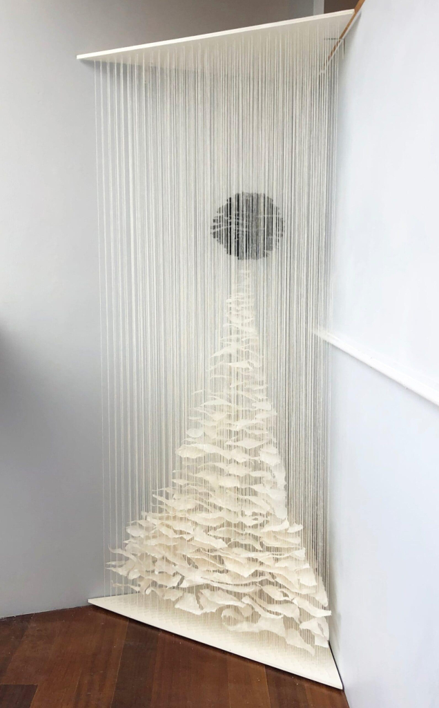 Sculpture installed in the corner of a gallery, made up of long cascading white threads with strips of white paper hanging from the bottom building up into a tall triangle, its point reaching a small black circle within the threads