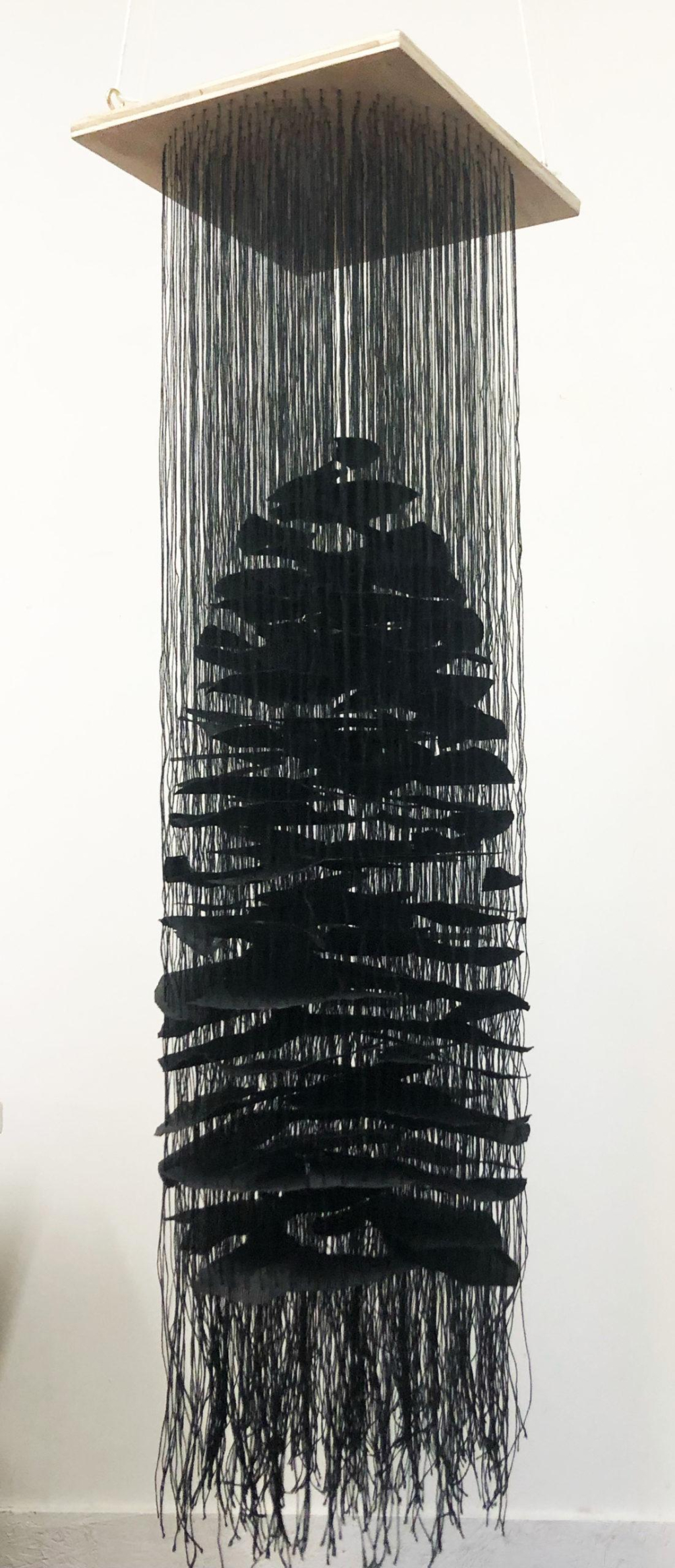 Hanging sculpture made up of long cascading black threads with strips of black paper arranged within, suspended from the ceiling so the threads are loose at the bottom