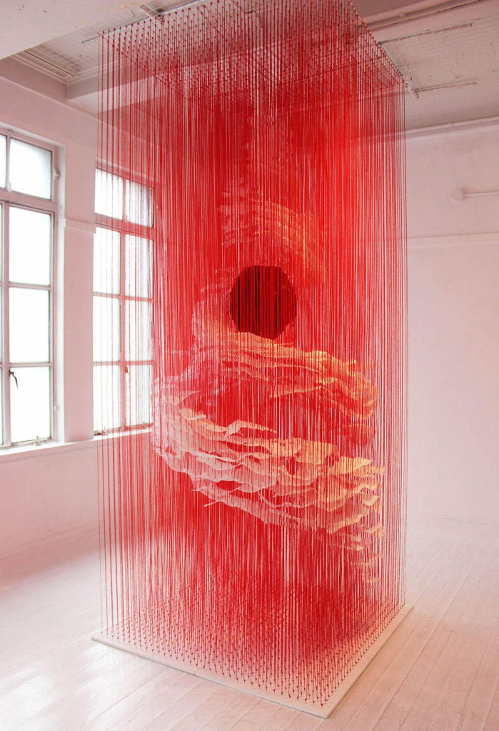 Hanging sculpture installed floor-to-ceiling in a gallery, made up of long cascading red threads with strips of white paper hanging within, arranged in swirling shapes, and a dark circle in the center