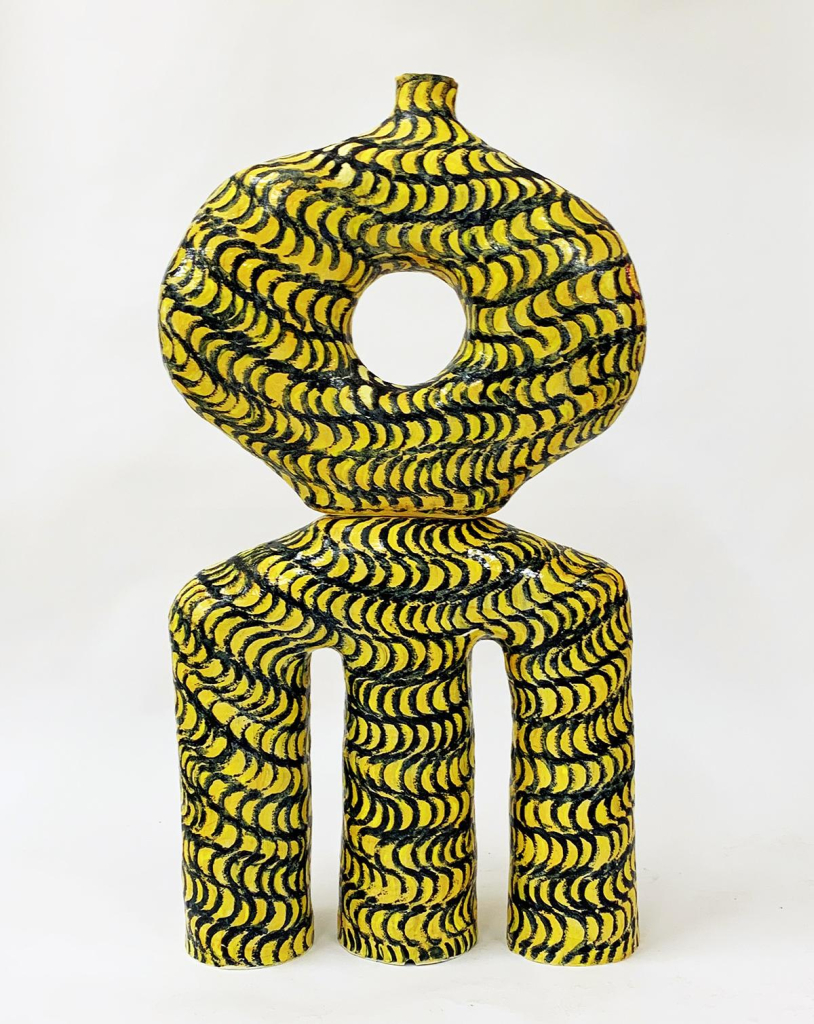 Abstract ceramic sculpture standing on three round legs with a big "O" shape on top, painted in a squiggly line pattern in black and yellow