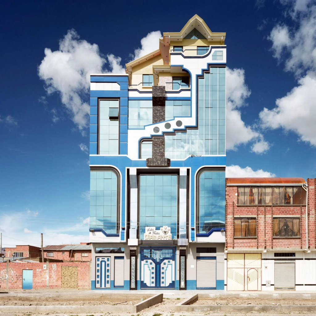 A narrow vertical building with bold geometric facade in white and blue colors. Playful geometric motifs run up and down the walls and the square glass windows reflect the blue and clouds of the sky.