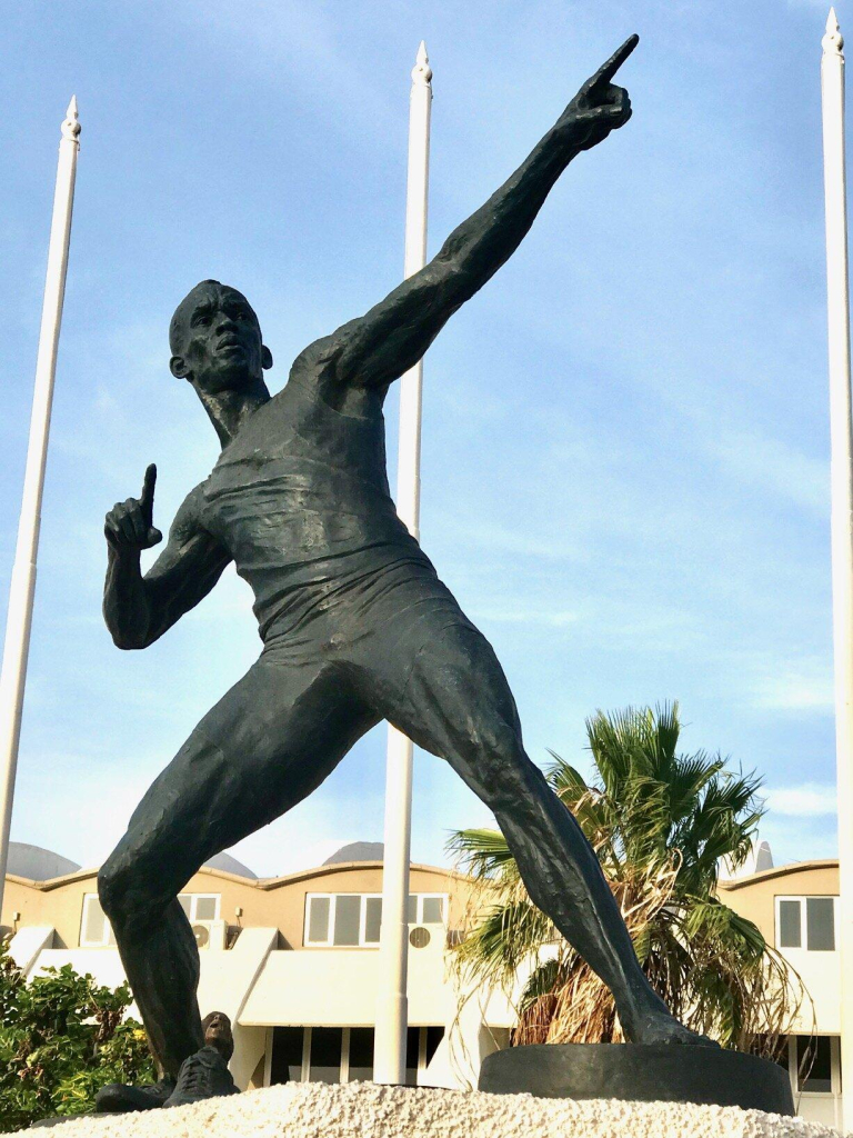 Bronze sculpture of a male figure standing with legs apart, leaning back and raising one arm straight up and the other bent, both pointing their index fingers