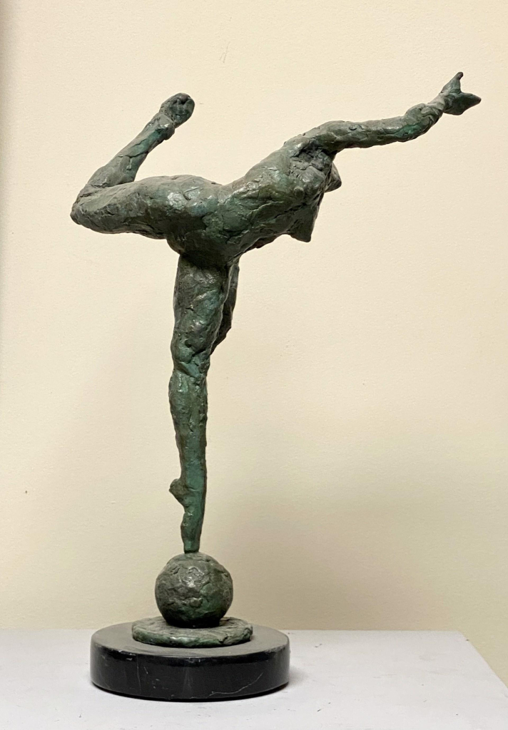 Bronze sculpture of a nude female figure balancing on her toes on top of a ball, her right leg bent back behind her and her body leaning forward, right arm oustretched