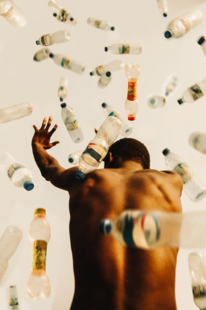 Photo of a shirtless Black man seen from the back surrounded by falling plastic bottles, with his arms outstretched as if trying to ward them off