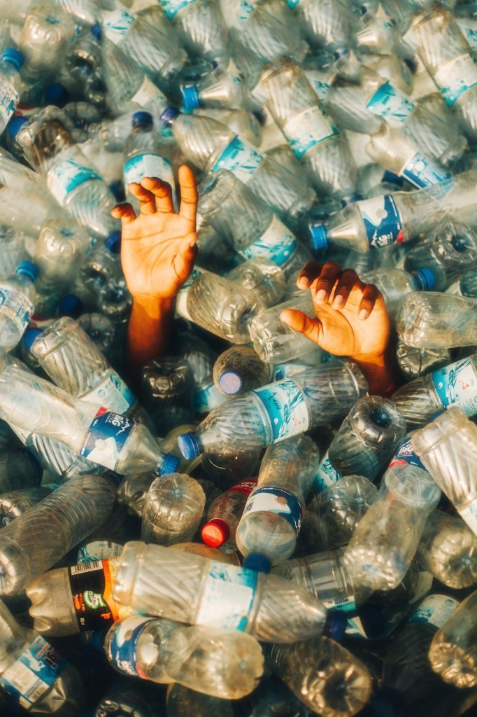 Photo of a large pile of plastic bottles filling the entire frame, with the outstretched hands of a Black figure reaching out from within, their body lost in the bottles