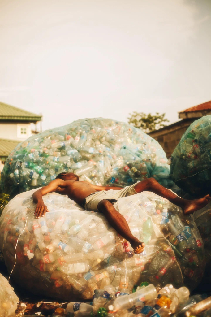 Photo of a shirtless Black man lying as if sleeping on a huge translucent bag full of plastic bottles, with other large bags and piles of bottles surrounding him