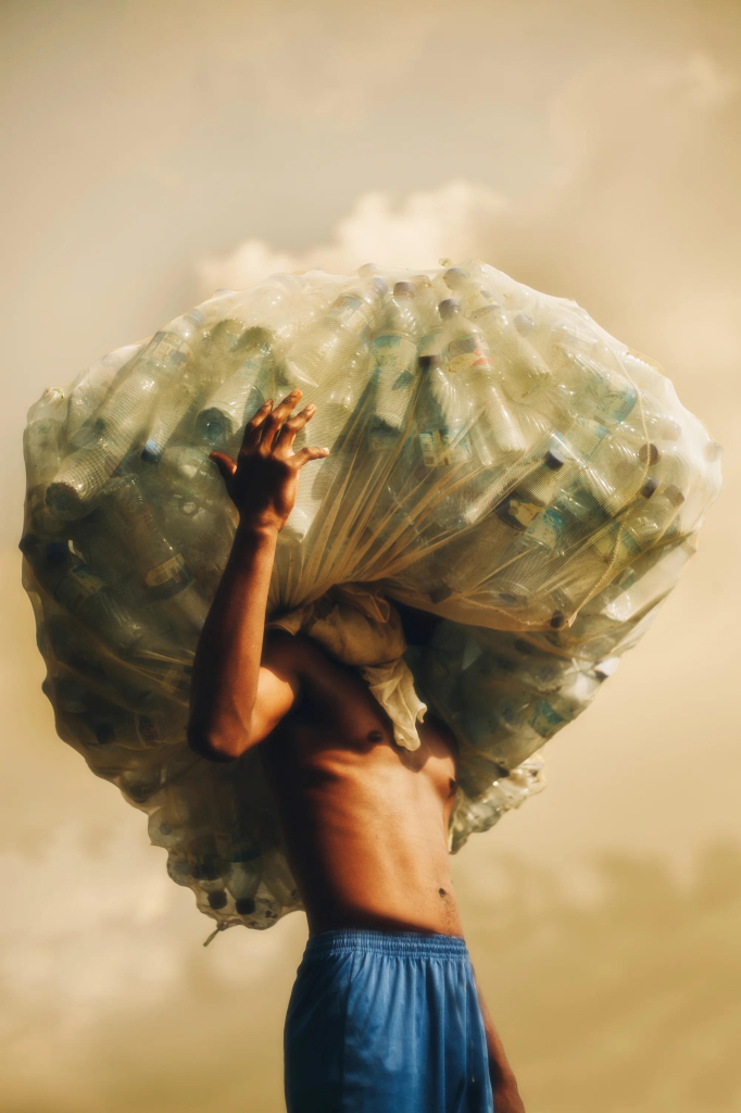 Photo of a shirtless Black man standing against a dusty white-brown sky, holding a huge translucent bag of plastic bottles on his head, totally obscuring his face