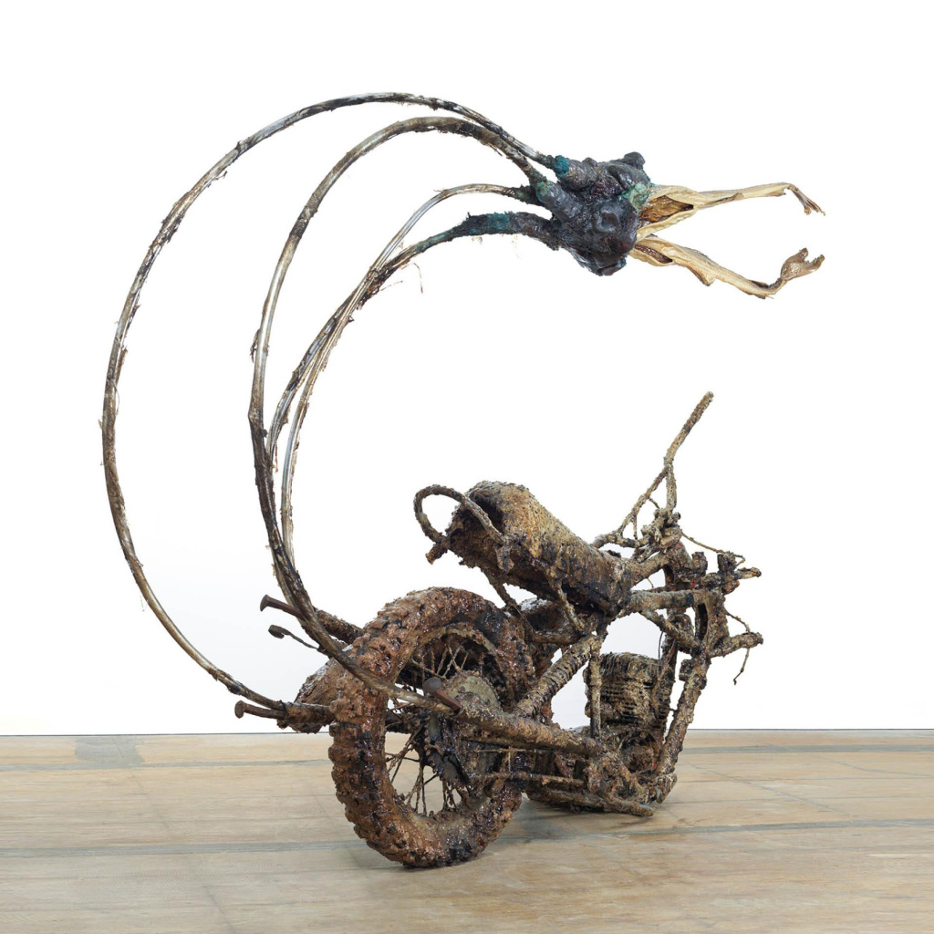 Sculpture of a partial section of a motorcycle with long tubes curving upward holding a beak-like form, all covered with a textured, rusty finish