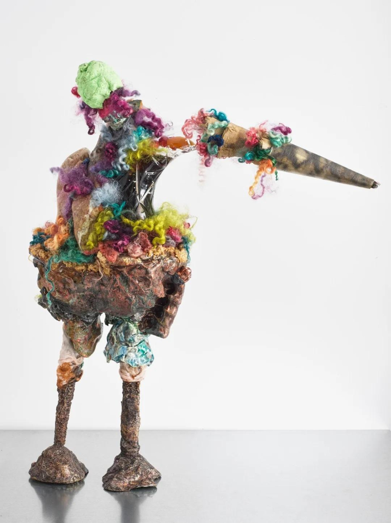 Sculpture of a standing bird-like figure made up of colorful bits of fuzzy material and ceramic elements