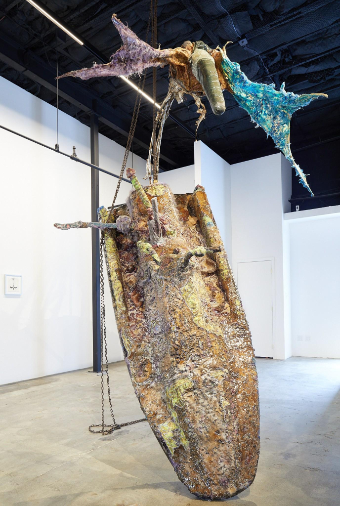 Sculpture of a decayed-looking jetski standing upward, above it is a birdlike figure made of found materials with wings outstretched