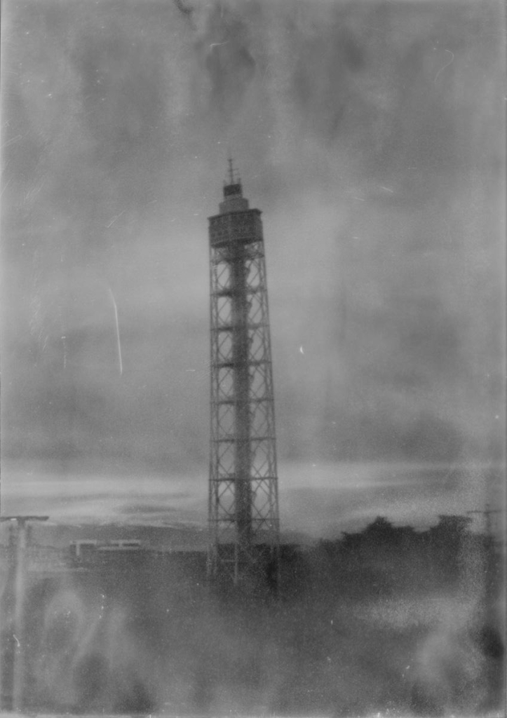 Slightly blurry black and white image of a narrow radio tower standing in a sparse, rural landscape