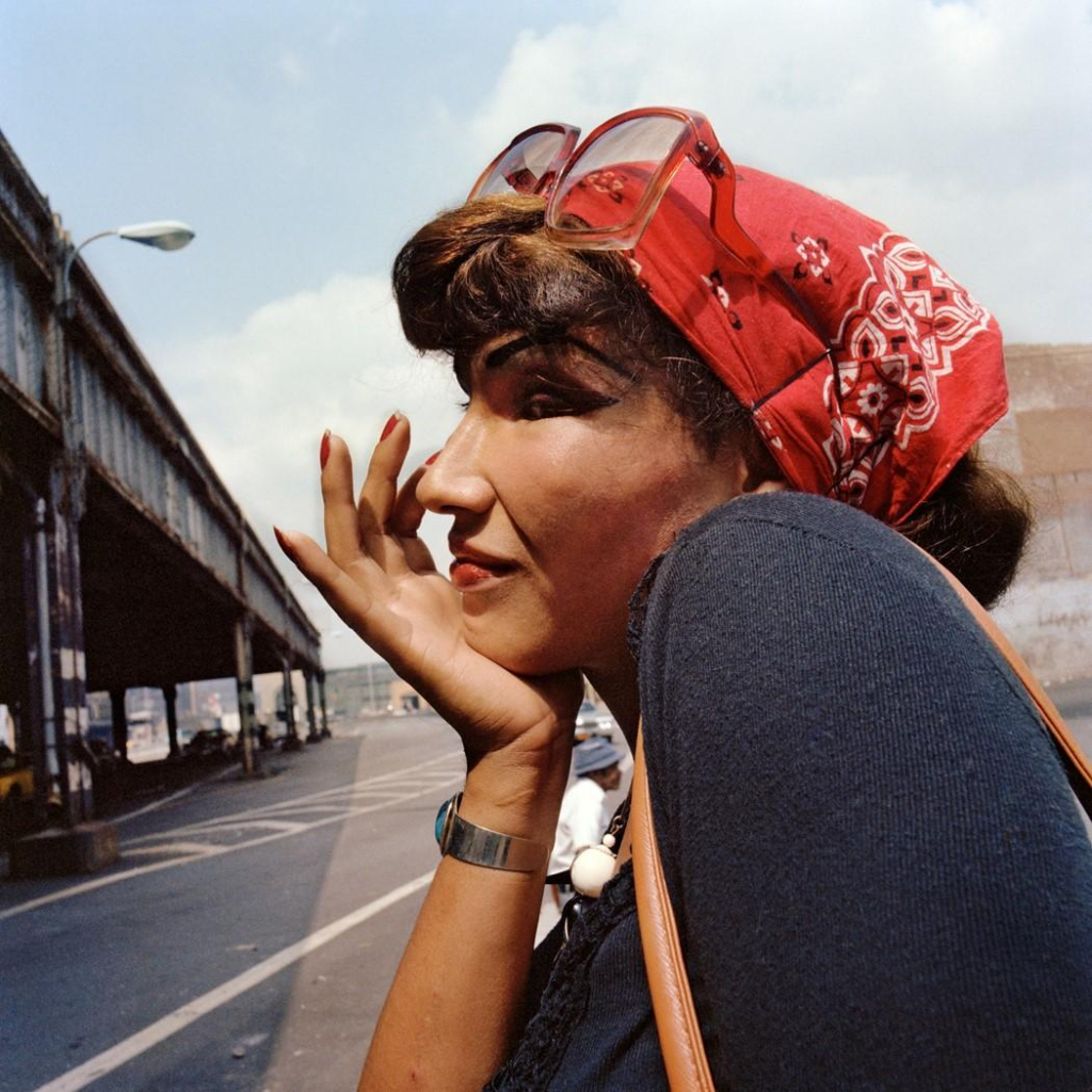 Photo of a woman with dark hair and heavy makeup shown in profile with her hand on her chin with a slight smile, wearing a red kerchief and large glasses on her head. Behind her is a city street and blue sky