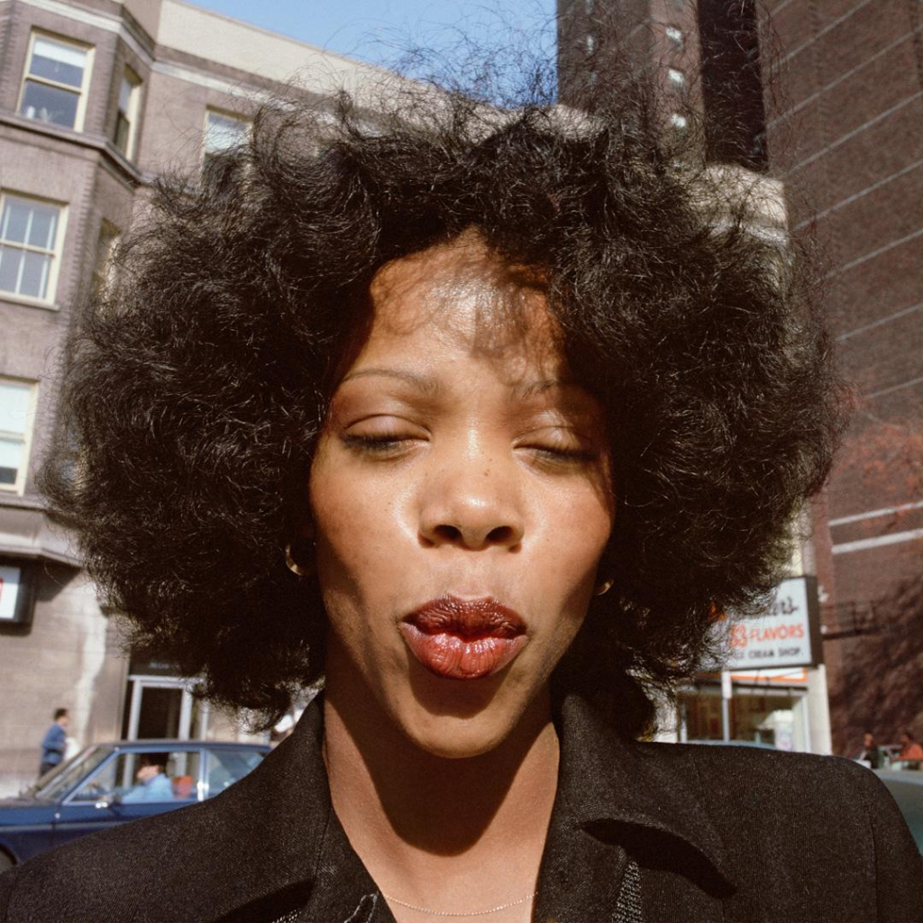 Photo of a Black woman with wavy hair and a black coat, pouting her lips with partially-closed eyes looking down at the camera. Behind her are city buildings and a street.