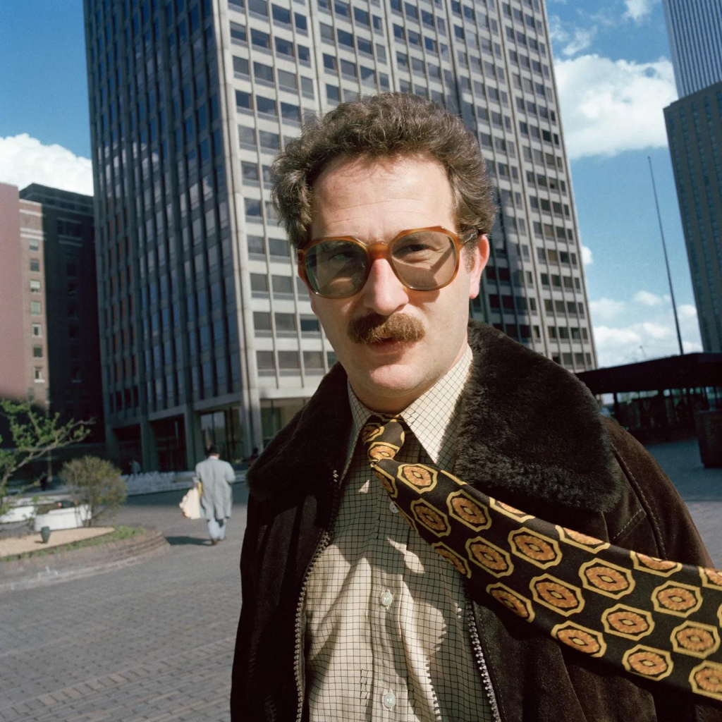 Photo of a white man with a mustache and tinted glasses, wearing a coat with a fur collar and a patterned tie blowing in the wind as he looks directly at the camera. City skyscrapers and a wide street are visible behind him