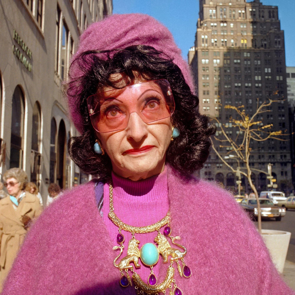 Photo of an older woman with dark hair and heavy makeup wearing a bright pink ensemble (including hat) with large gold jewelry and large pink glasses. She looks just above the camera with a kind of grimace. City skyscrapers are visible behind her