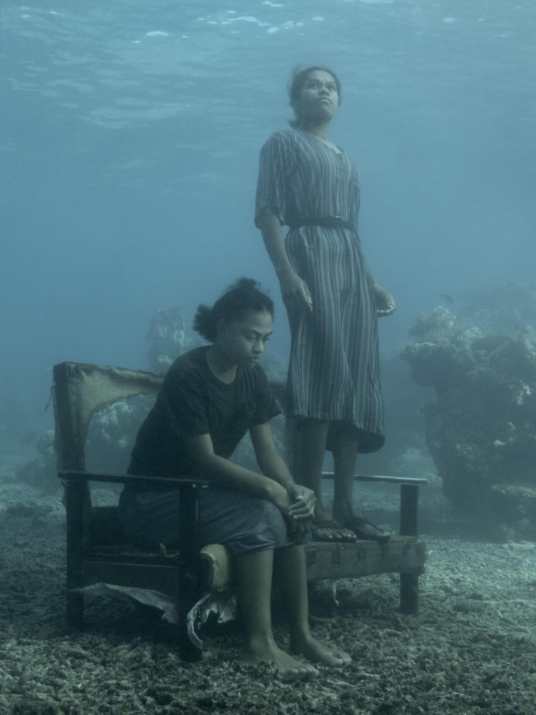 Underwater photo of two women, one standing on a decaying cough and the other sitting with her hands on her knees