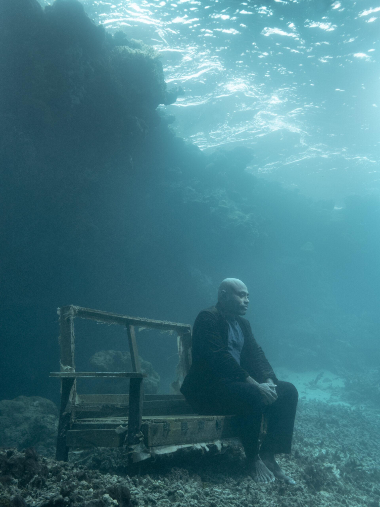 Underwater photo of a man in a suit sitting on a decaying bench, hands folded in his lap