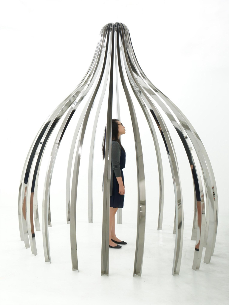 A woman standing within a large metal sculpture with a rounded base that comes to a point at the top, made up of mirrored legs repeating around in a circle